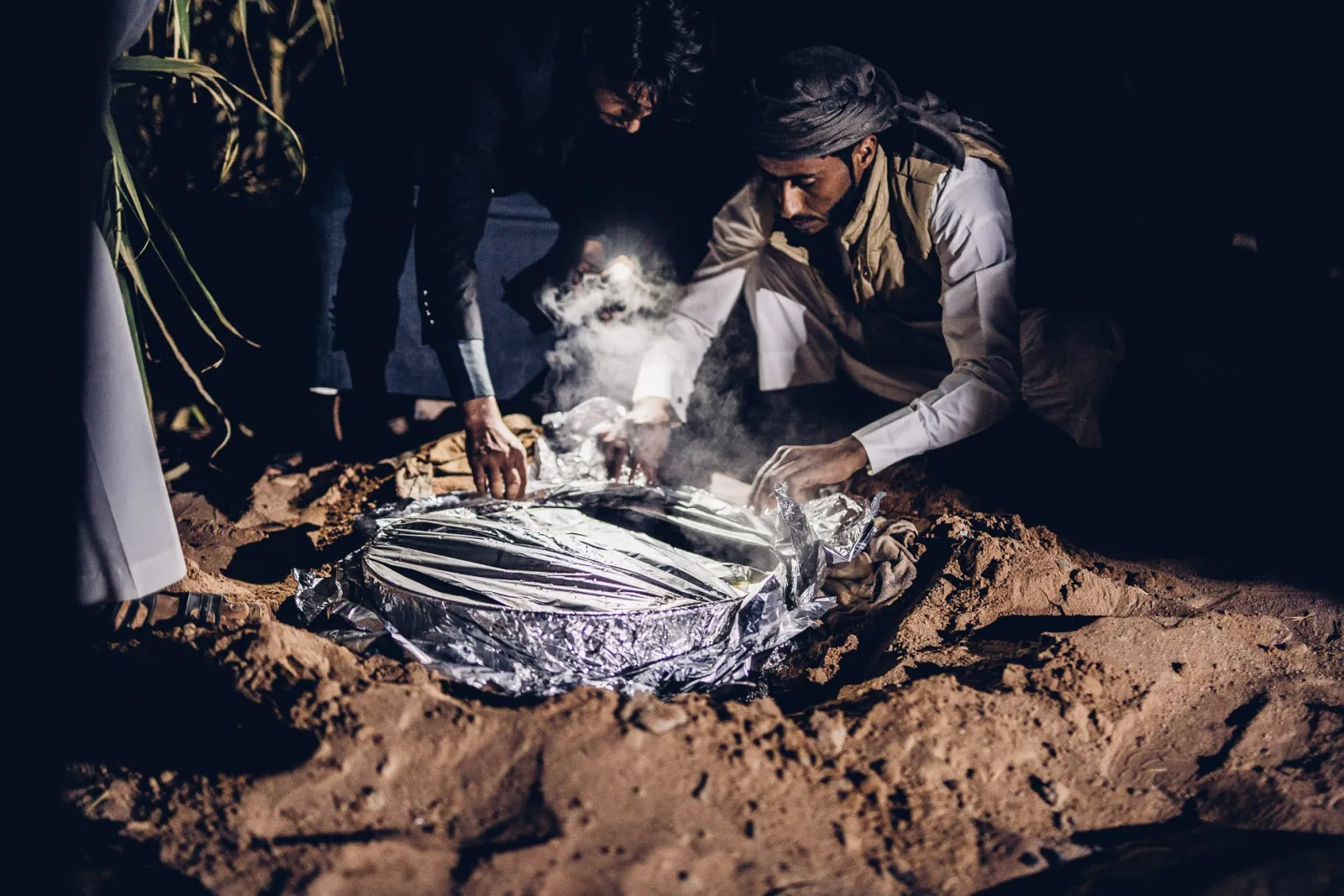 Bedouin hosts lifting a foil-wrapped zarb dinner out of the sand after dark