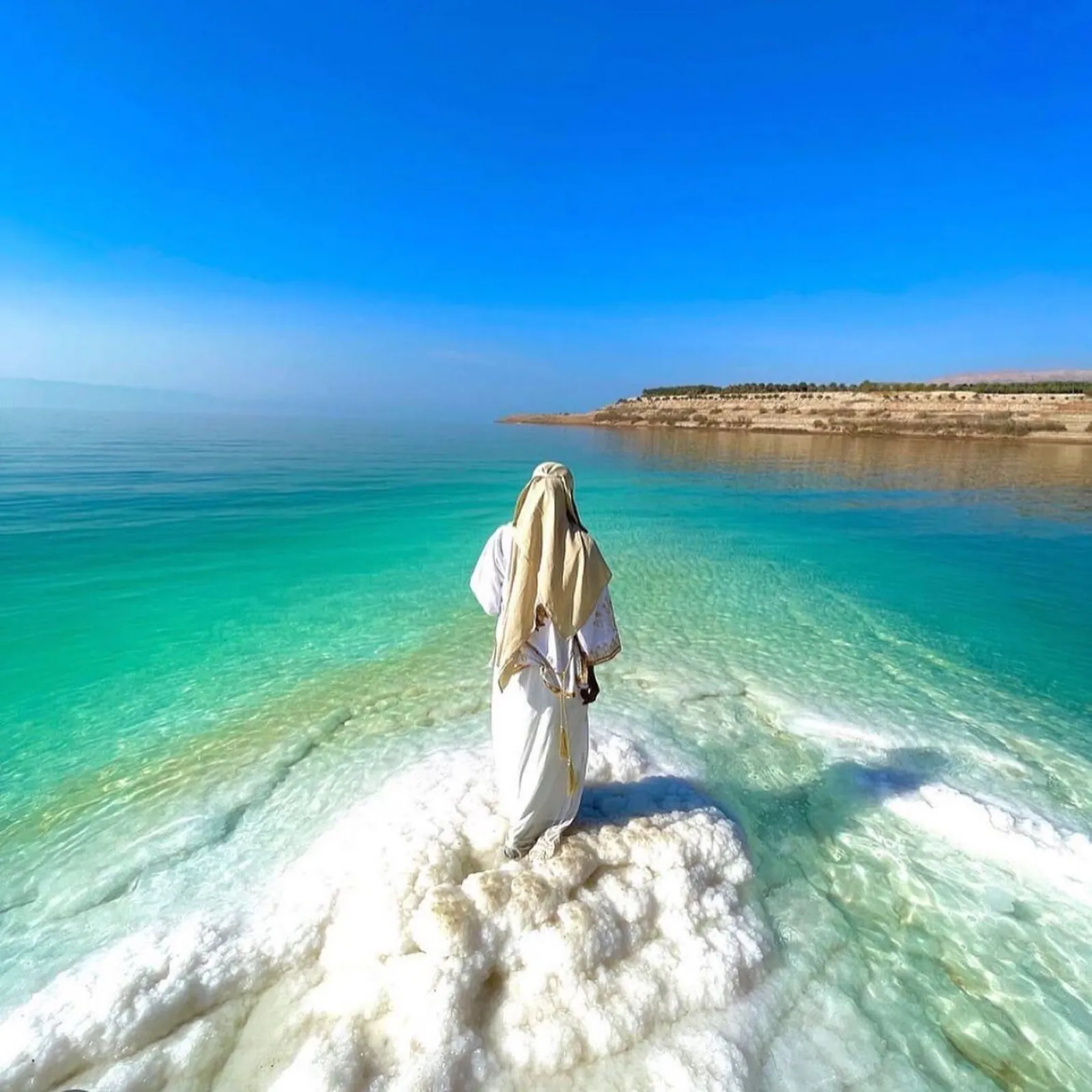 A figure standing on a white salt platform at the edge of the Dead Sea